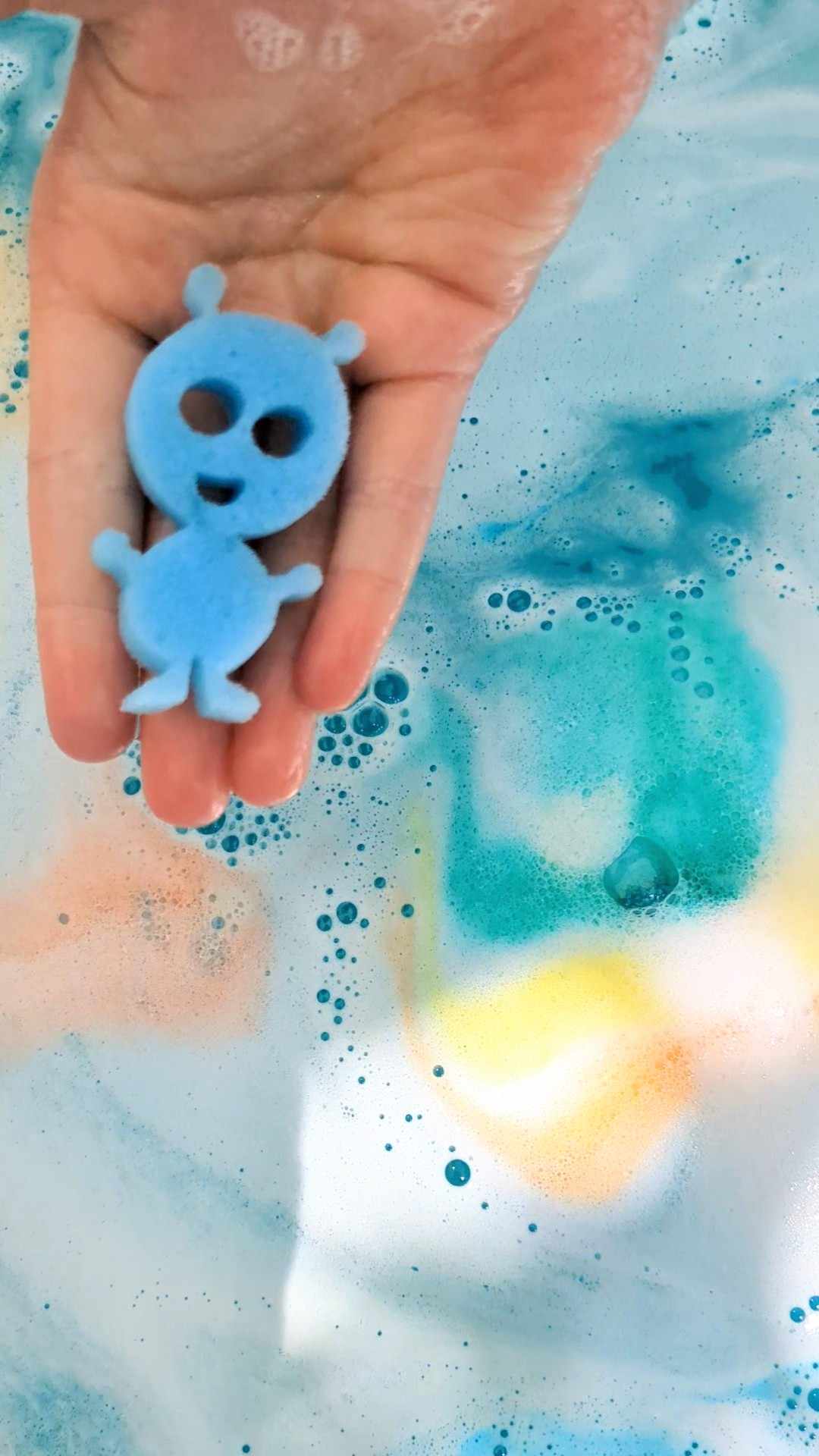 Child's hand holding a blue space-themed sponge toy over rainbow bath water