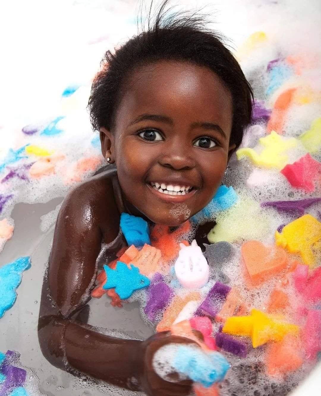 Smiling girl in the bath surrounded by colourful sponge toys