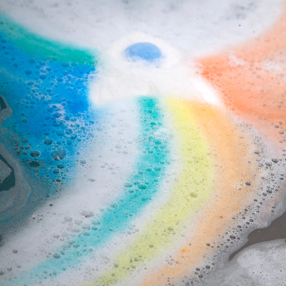 Rainbow bath bomb fizzing in white foam with blue, yellow and orange swirls