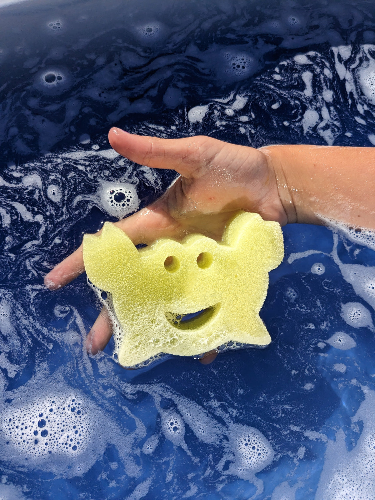 Child's hand holding a yellow crab sponge toy in dark blue bath water