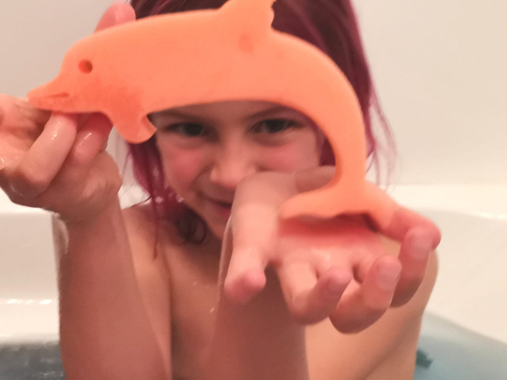 Girl in the bath holding an orange dolphin sponge toy from a Jumbo Bath Bomb Sprudel