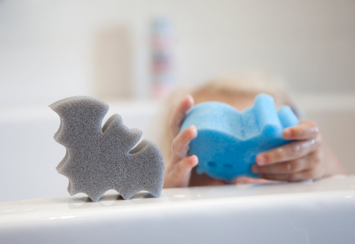 big bat-shaped sponge and blue round sponge held by a childs hand on a white bath tub