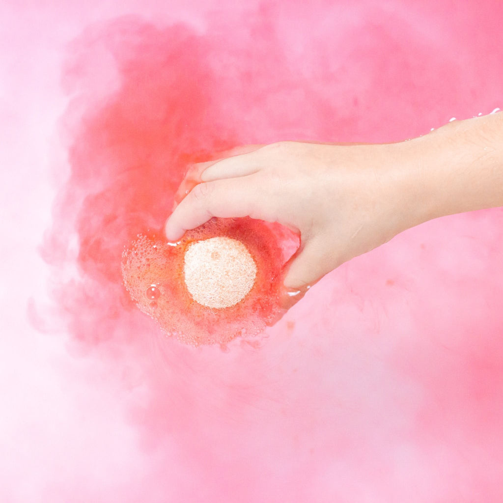 Child's hand holding a coral kids bath bomb fizzing in pink bath water