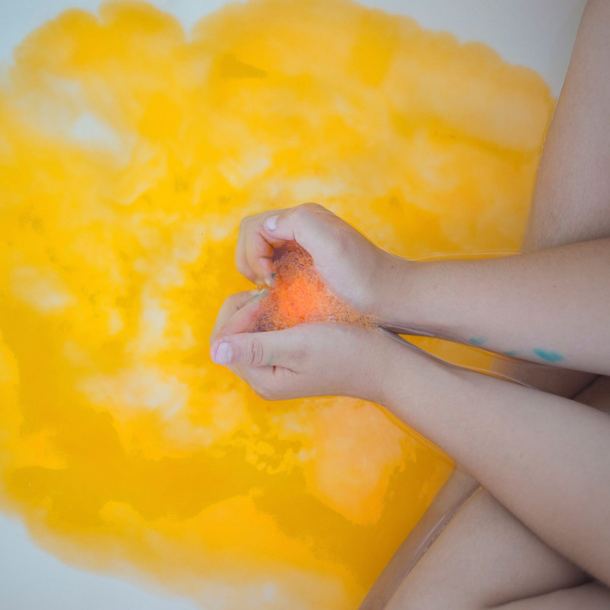 A close-up of childs hands holding fizzing orange bath bomb with bubbles, with bright yellow-orange bath water filling the background.